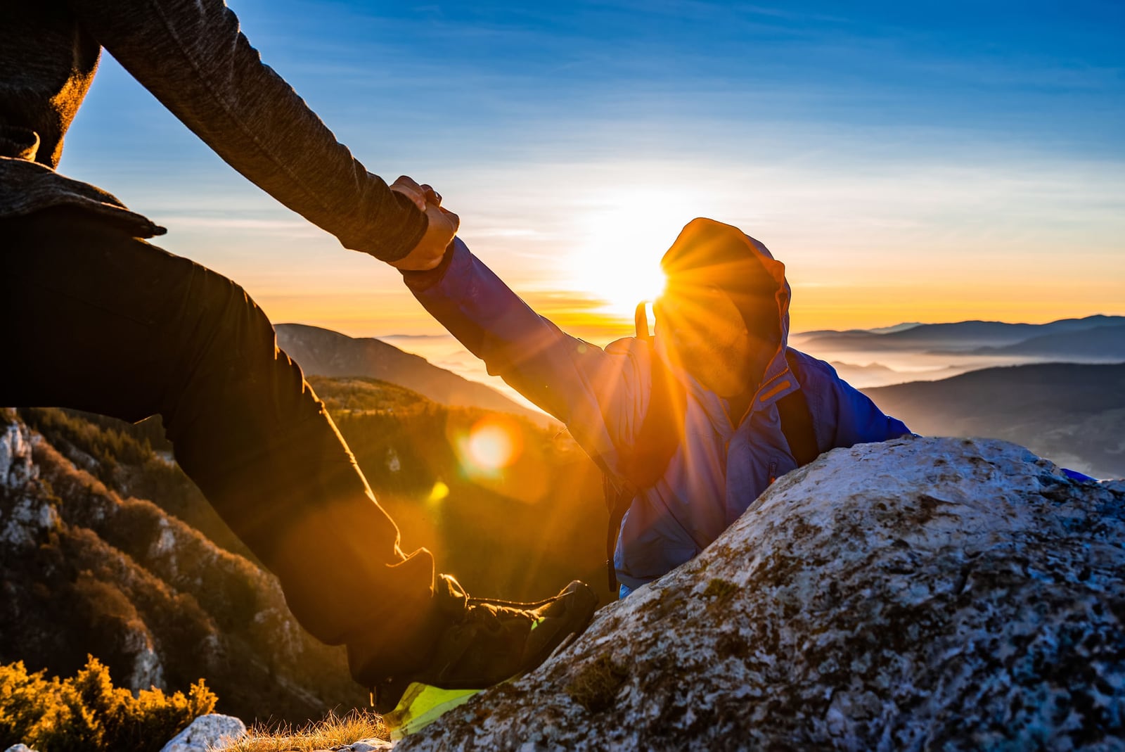 Eine helfende Hand zieht eine Person beim Sonnenaufgang auf den Gipfel — Sinnbild für „Moving Mountains for CARE“